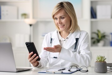 Smiling doctor with smartphone having online consultation at table in office