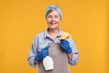 Happy housewife with spray bottle and rag on orange background