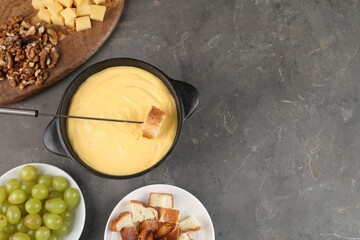 Fondue pot with tasty melted cheese, fork and different snacks on grey table, flat lay. Space for...