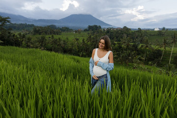 Pregnant woman wearing  casual clothes  holding her belly outside with newly planted rice field and cloudy sky in background