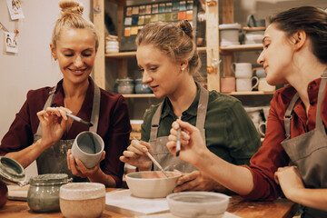 A company of three young women friends are painting ceramics in a pottery workshop.