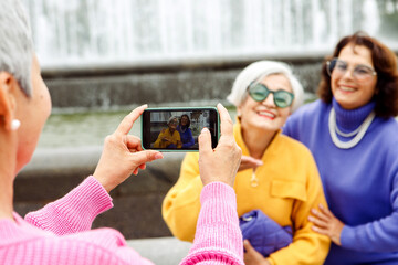 Three elderly friends in bright sweaters take a photo on a smartphone and laugh, having a great...