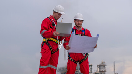 Engineer wear uniform and helmet stand workplace hand holding tablet computer, survey inspection team work see detail blue print plant site to work with oil refinery  background. - 800795659