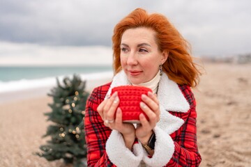 Lady in plaid shirt with a red mug in her hands enjoys beach with Christmas tree. Coastal area. Christmas, New Year holidays concep