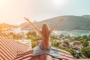 Woman sits on rooftop with outstretched arms, enjoys town view and sea mountains. Peaceful rooftop...