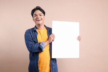 Friendly Young Man In Casual Style Smiling and Showing Blank White Board for Advertisement Isolated on Beige Background