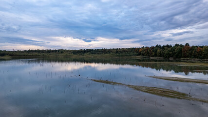 The image conveys a moment of tranquility at dusk with a view across a calm lake reflecting the evening sky. The horizon is lined with a forest showing early signs of autumnal color, adding a touch of