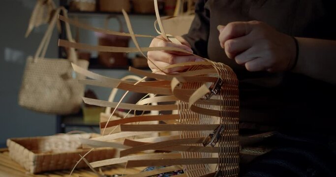Slow motion scene while a beautiful Asian female artisan who is a local villager weaving folk basket made from leftover paper band to sell as extra income in the basketry shop shop.