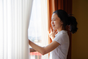 Asian woman waking up after sleeping standing up from bed and opening curtains looking at window at home in sunny morning.