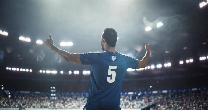 Excited Footballer with His Back Turned to Camera Standing In Front of a Stadium with Fans, Showing Thumbs Up and Warming Up the Crowd. Young Man in Blue Uniform Getting Psyched Up Before a Game