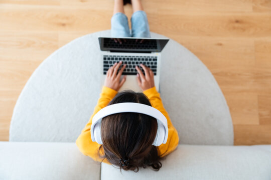 Top view of young Asian woman sitting on the floor and working on computer laptop, wearing wireless headset, listening to music