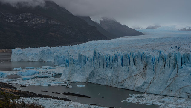 An amazing glacier stretches between the mountain slopes to the horizon. A mass of blue ice with cracks, sharp peaks, and crevices. Thawed ice floes float in a glacial lake. Cloudy. Fog. Perito Moreno