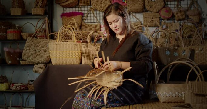 Slow motion scene while a beautiful Asian female artisan who is a local villager weaving folk basket made from leftover paper band to sell as extra income in the basketry shop shop.