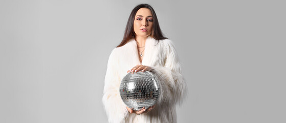 Fashionable beautiful young woman in fur coat with disco ball on grey background