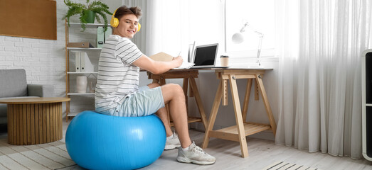 Young man in headphones on fitball using laptop at home