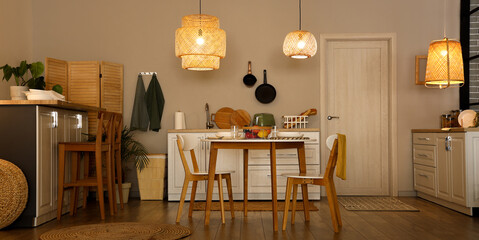 Interior of modern kitchen with glowing lamps, dining table and white counters at evening