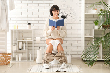 Young woman reading book on toilet bowl at home