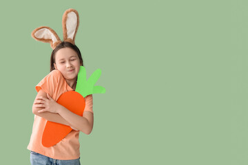 Happy little girl in Easter bunny ears with paper carrot on green background