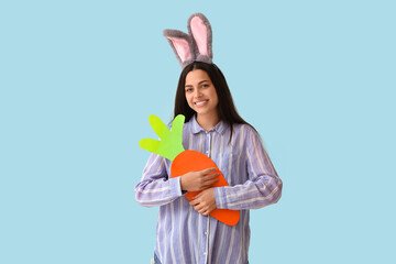 Beautiful young happy woman in Easter bunny ears with paper carrot on blue background
