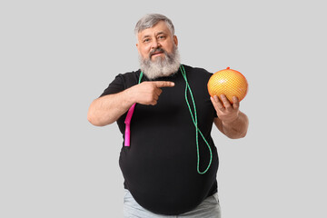 Overweight mature man with jumping rope pointing at pomelo fruit on grey background. Weight loss...