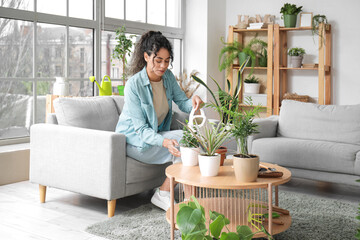 Beautiful young African-American woman watering plants sitting on sofa in living room