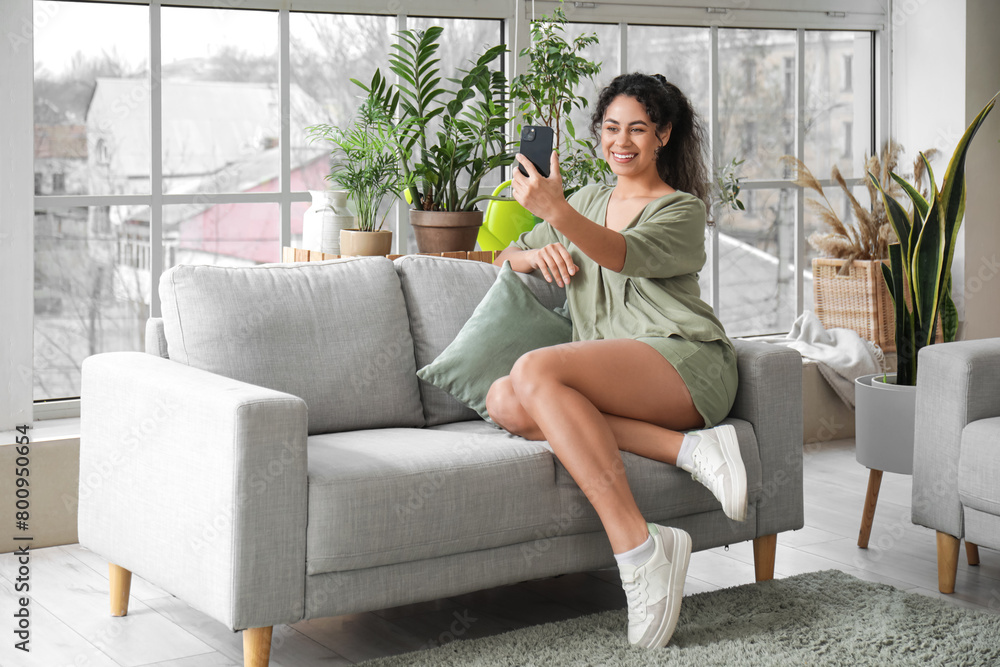 Poster beautiful young african-american woman taking selfie with plants in living room