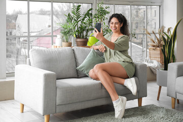 Fototapeta premium Beautiful young African-American woman taking selfie with plants in living room