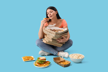 Upset young woman with unhealthy food on blue background. Overeating concept