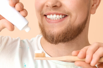 Smiling young bearded man pouring tooth paste onto brush on beige background, closeup