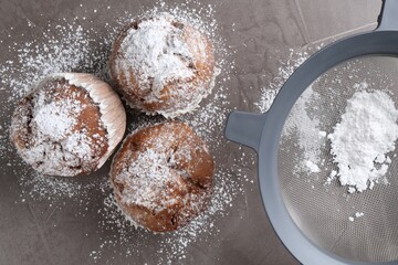 Sieve with sugar powder and muffins on grey textured table, flat lay