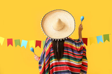 Young woman in Mexican sombrero hat and poncho holding maracas on yellow background, back view