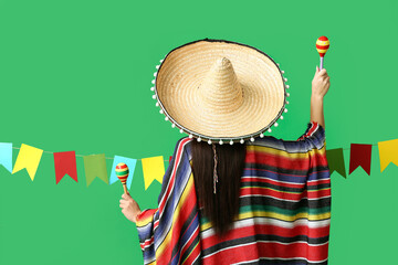 Young woman in Mexican sombrero hat and poncho holding maracas on green background, back view