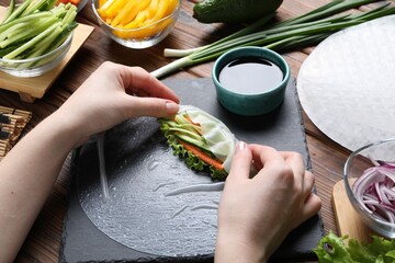 Woman wrapping spring roll at wooden table with products, closeup
