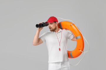 Young male lifeguard with ring buoy looking through binoculars on grey background