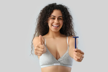 Beautiful young happy woman with razor showing thumb-up gesture on grey background. Depilation...