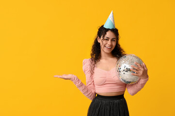 Beautiful young African-American woman in party hat with disco ball pointing at something on yellow...
