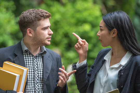 A man and a woman are arguing in a park. The woman is pointing her finger at the man while the man is holding books and gesturing with his hands.