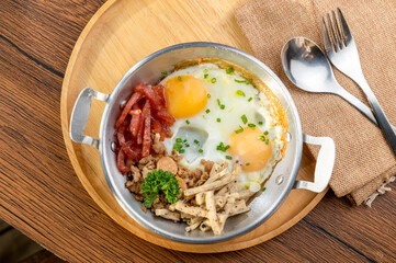A photo of a dish of food that has two eggs, bacon, and other ingredients in a metal pan on a wooden table.
