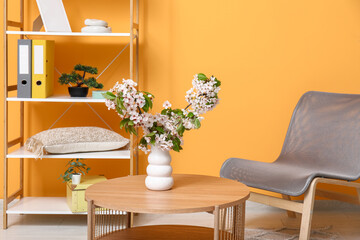 Vase with blossoming branches on coffee table near orange wall in living room