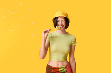 Teenage girl with sweet lollipop and soap bubbles on yellow background