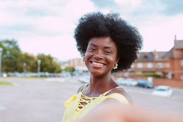 Portrait of a young cheerful black afro woman taking selfie at city park. Wears yellow top and...