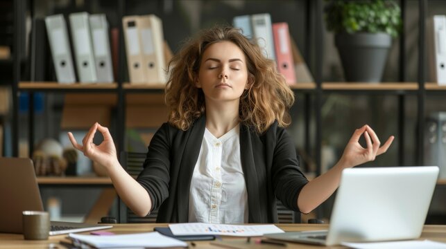 Calm young executive meditating at her desk with laptops and documents around, balancing stress with serenity