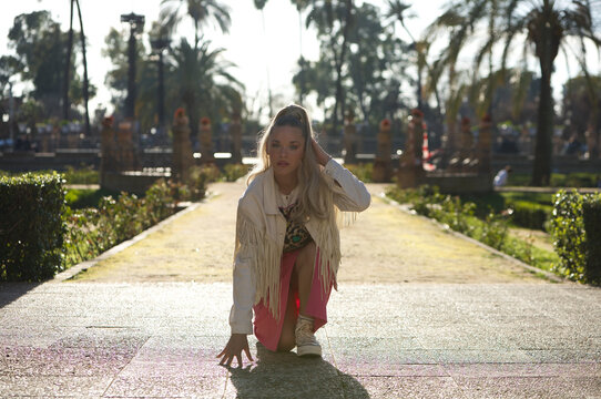 Young beautiful blonde woman with a ponytail in her hair wearing a pink skirt and fringed jacket rests one knee, one hand on the ground and with a defiant face looks at the camera. The girl is poised.