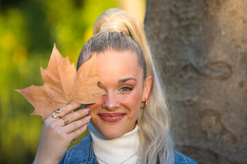 Pretty young blonde woman with a ponytail in her hair covers one eye with a dry leaf. Focus on the...