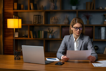 Young successful employee business woman sitting  at workplace in suit and hold paper account documents with laptop in modern office