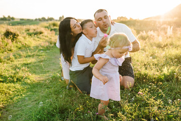 Mother, father and children playing. Happy family in evening sunlight. Daughter, son, parents walk on green and yellow grass and blowing soap bubbles in spring field at sunset. Concept summer holiday.