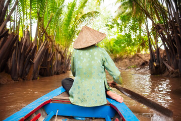 Woman with asian hat rowing a boat by coconut palm trees at Mekong delta