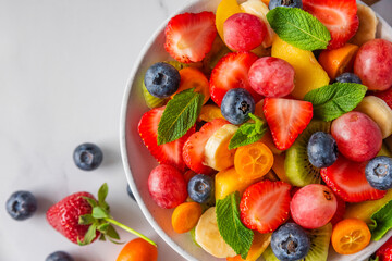 Summer fresh fruit salad in a bowl on white marble background. Top view. Healthy food for...