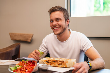 guy with shawarma. a cheerful young guy in a white T-shirt sits at a table against the background of a window and eats a large shawarma, fast food concept