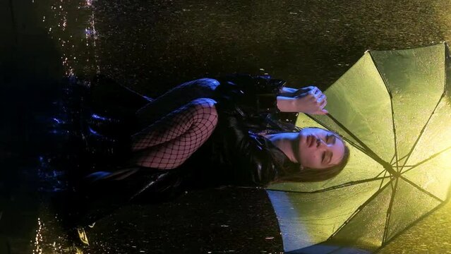 Woman in squats beneath parasol lit by street lamp. Frustrated young lady in dimly surrounding covers up with umbrella from downpour